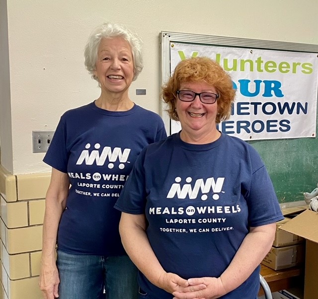 Two Meals on Wheels volunteers in matching shirts standing beside a Volunteers Are Our Hometown Heroes banner